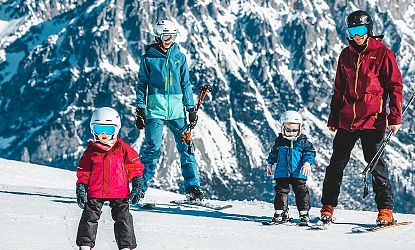 Eine Familie beim Skifahren auf einer verschneiten Piste. Mutter, Vater und zwei Kinder tragen bunte Skibekleidung und Helme vor einer bergigen Winterkulisse.