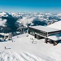 Bergstation mit breiter Skipiste und einzelnen Skifahrern auf einem verschneiten Gipfel, dahinter weites Panorama der winterlichen tiroler Alpen.