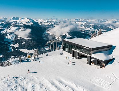 Bergstation mit breiter Skipiste und einzelnen Skifahrern auf einem verschneiten Gipfel, dahinter weites Panorama der winterlichen tiroler Alpen.