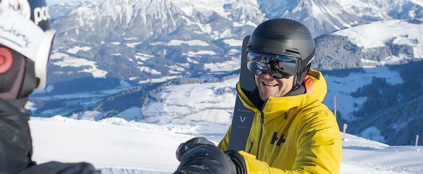 Mann in gelber Jacke und Helm genießt einen sonnigen Tag beim Skifahren in den verschneiten Bergen mit malerischem Panorama im Hintergrund.