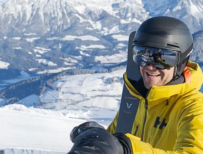 Mann in gelber Jacke und Helm genießt einen sonnigen Tag beim Skifahren in den verschneiten Bergen mit malerischem Panorama im Hintergrund.