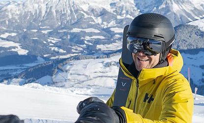 Mann in gelber Jacke und Helm genießt einen sonnigen Tag beim Skifahren in den verschneiten Bergen mit malerischem Panorama im Hintergrund.