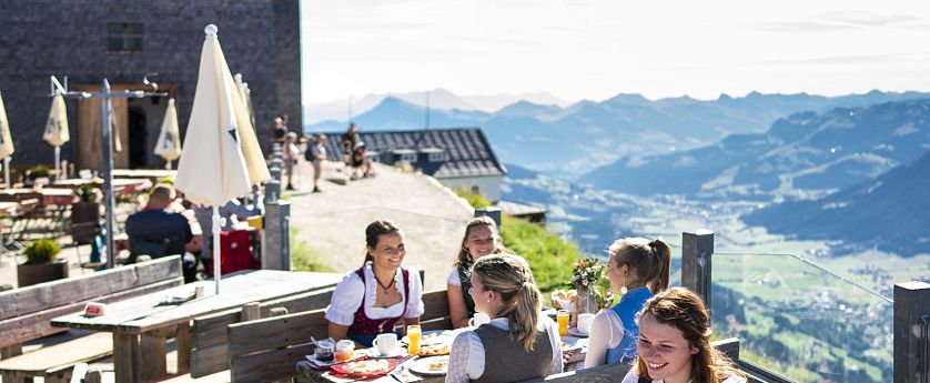 Menschen in traditioneller Tracht genießen ein ausgiebiges Frühstück auf der sonnigen Panoramaterrasse der Hohen Salve Gipfelalm.