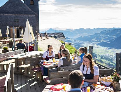 Menschen in traditioneller Tracht genießen ein ausgiebiges Frühstück auf der sonnigen Panoramaterrasse der Hohen Salve Gipfelalm.