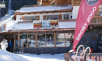 Das Bild zeigt ein Sportgeschäft in den Bergen, umgeben von Schnee. Ein großes Schild weist auf Ski- und Rodelverleih sowie einen Ausverkauf hin. Im Vordergrund stehen Skiausrüstungen.
