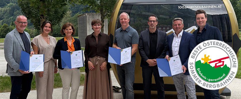 Sechs Personen stehen lächelnd zusammen in einem Freiluftbereich. Sie halten Zertifikate in den Händen, im Hintergrund ist eine Seilbahn zu sehen.