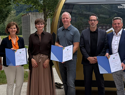 Sechs Personen stehen lächelnd zusammen in einem Freiluftbereich. Sie halten Zertifikate in den Händen, im Hintergrund ist eine Seilbahn zu sehen.