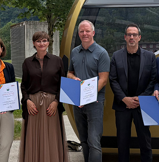 Sechs Personen stehen lächelnd zusammen in einem Freiluftbereich. Sie halten Zertifikate in den Händen, im Hintergrund ist eine Seilbahn zu sehen.