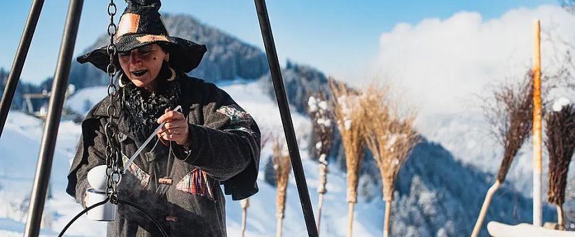 Person in Hexenkostüm bei Schneelandschaft, kocht über Feuerstelle. Hintergrund zeigt schneebedeckte Berge und klare Himmel.