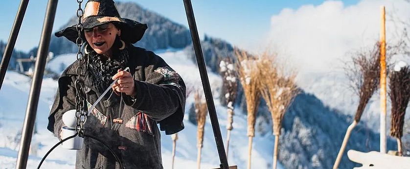 Eine Person in einem schwarzen Umhang und einer Hexen-Hut steht vor einem großen Kessel, während sie mit einem Löffel rührt. Die schneebedeckte Landschaft und Fellen im Hintergrund schaffen eine winterliche Atmosphäre.
