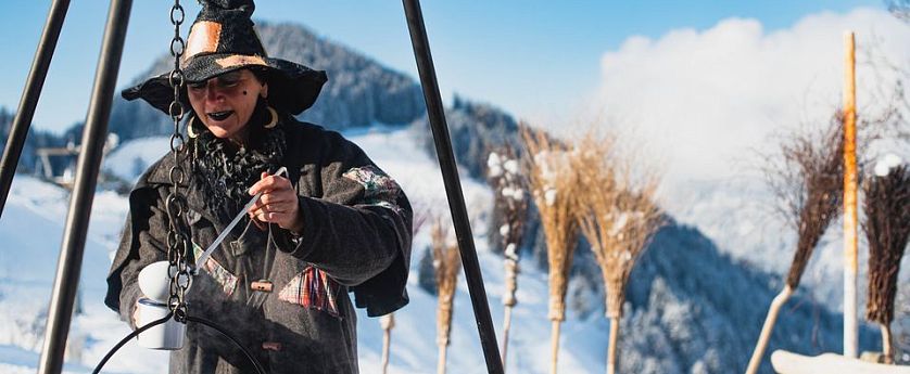 Persona disfrazada de brujo sirviendo de un caldero en un evento invernal en un paisaje montañoso nevado con escobas alineadas.