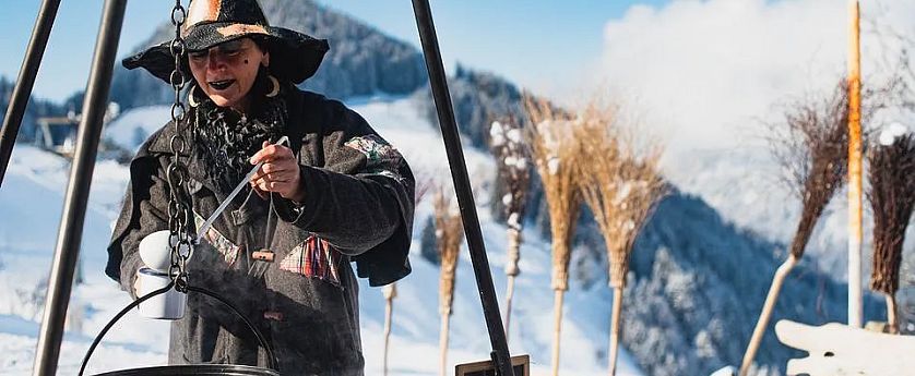 Een persoon in een heksenkostuum staat buiten in een besneeuwd berglandschap, terwijl ze een ketel roeren. Houten bezems staan op de achtergrond.