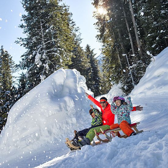 Familie rodelt auf der Rodelbahn Hexenritt in Söll mit gehobenen Armen und fröhlichen Gesichtern