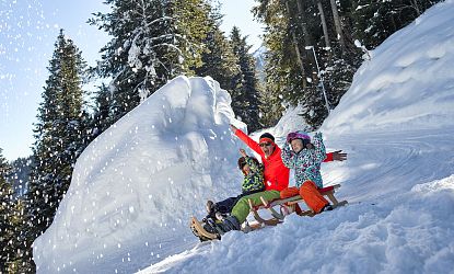 Familie rodelt auf der Rodelbahn Hexenritt in Söll mit gehobenen Armen und fröhlichen Gesichtern