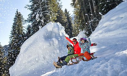 Familie rodelt auf der Rodelbahn Hexenritt in Söll mit gehobenen Armen und fröhlichen Gesichtern