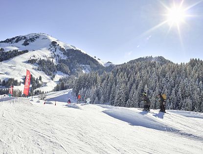 Verschneite Skipiste mit Funpark-Elementen und roten Fahnen im Skigebiet Söll, Kinder beim Skifahren, im Hintergrund verschneite Bäume und ein sonniger Himmel