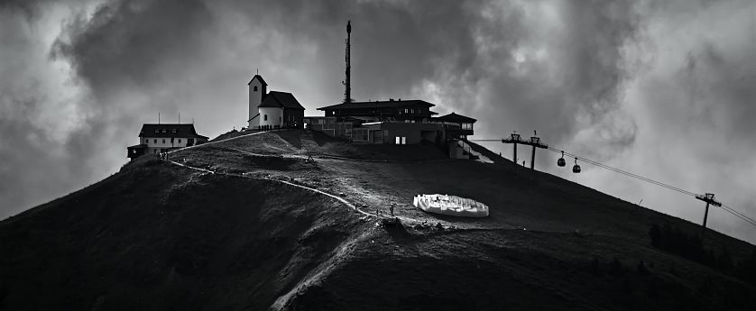 Schwarz-Weiß-Bild einer Berglandschaft mit einem kleinen Dorf, einem Aussichtsturm, einer Seilbahn und Wolken am Himmel.