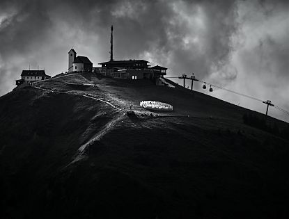 Schwarz-Weiß-Bild einer Berglandschaft mit einem kleinen Dorf, einem Aussichtsturm, einer Seilbahn und Wolken am Himmel.