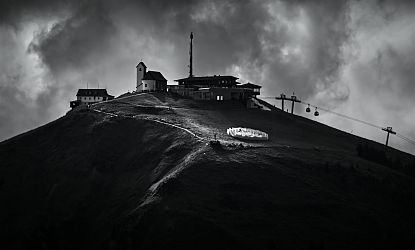 Schwarz-Weiß-Bild einer Berglandschaft mit einem kleinen Dorf, einem Aussichtsturm, einer Seilbahn und Wolken am Himmel.