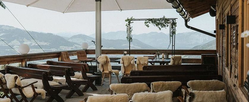 Die Terrasse eines Alpengasthauses ist mit Holzstühlen und Schaffellen ausgestattet. Blick auf die Berge, mit einem Pavillon in der Mitte, der von Pflanzen dekoriert ist.