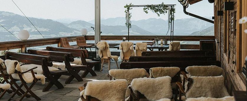 Die Terrasse eines Alpengasthauses ist mit Holzstühlen und Schaffellen ausgestattet. Blick auf die Berge, mit einem Pavillon in der Mitte, der von Pflanzen dekoriert ist.