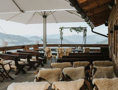 Die Terrasse eines Alpengasthauses ist mit Holzstühlen und Schaffellen ausgestattet. Blick auf die Berge, mit einem Pavillon in der Mitte, der von Pflanzen dekoriert ist.