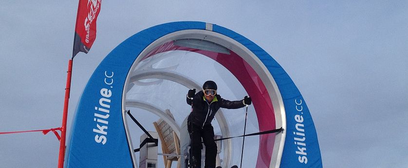 Skifahrer am Start einer Skiline-Rennstrecke, bereit zur Abfahrt durch ein rundes Starttor mit blau-weiß-roter Umrandung und Beschriftung „skiline.cc