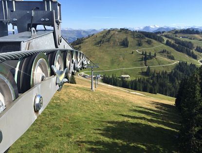 Berglandschaft mit Seilbahnrollen im Vordergrund; sanfte Hügel und schneebedeckte Berge im Hintergrund unter klarem Himmel.