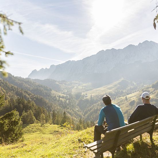 Zwei Wanderer sitzen auf einer Holzbank und genießen den Ausblick über ein sonnendurchflutetes Tal mit bewaldeten Hügeln und der markanten Silhouette des Wilden Kaisers im Hintergrund