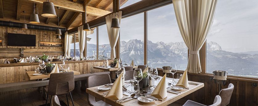 Das stilvoll gedecktes Bergrestaurant Kaiserlounge am Hartkaiser mit großen Panoramafenstern, durch die ein beeindruckender Blick auf das majestätische Kaisergebirge fällt. Die Einrichtung aus Holz schafft eine gemütlich-alpine Atmosphäre