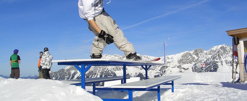 Ein Snowboarder führt einen Trick auf eine blau weißen Bank-Rail im Snowpark aus, mit schneebedeckten Bergen und blauem Himmel im Hintergrund