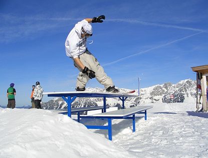 Ein Snowboarder führt einen Trick auf eine blau weißen Bank-Rail im Snowpark aus, mit schneebedeckten Bergen und blauem Himmel im Hintergrund
