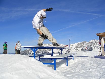 Ein Snowboarder führt einen Trick auf eine blau weißen Bank-Rail im Snowpark aus, mit schneebedeckten Bergen und blauem Himmel im Hintergrund