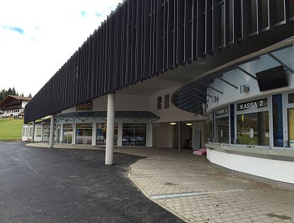 Eine moderne Gebäudeansicht mit glatten Linien und großen Fenstern, die zu einem Kassenbereich führt. Die Fassade ist teilweise aus schwarzem Material und steht im Kontrast zum Himmel.