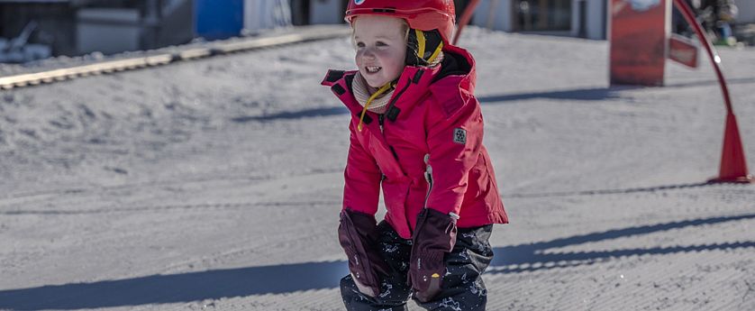 Kleines Kind in roter Skijacke und Helm fährt auf Skiern über eine präparierte Piste.