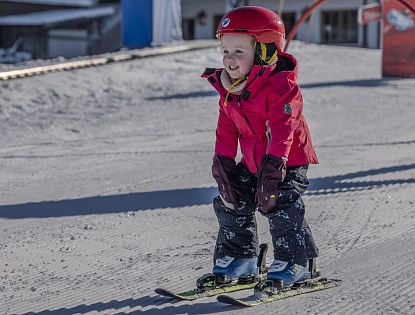 Kleines Kind in roter Skijacke und Helm fährt auf Skiern über eine präparierte Piste.