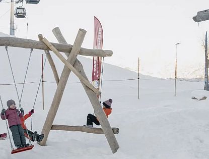 Winterlandschaft mit Holzspielplatz. Kinder schaukeln im Schnee, alle warm angezogen. Skilifte und schneebedeckte Berge im Hintergrund.