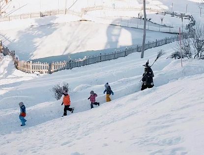 Fünf Kinder in bunter Winterkleidung laufen einen schneebedeckten Hang hinauf, geführt von einer Person in traditioneller Kleidung. Im Hintergrund ist ein umzäunter Bereich.