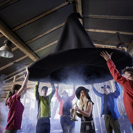 Gruppe von Menschen halten gemeinsam einen riesigen Hexenhut in einer rustikalen Hütte mit Nebeleffekten, während eine Frau mit Zauberhut und Trompete darunter steht und lächelt.
