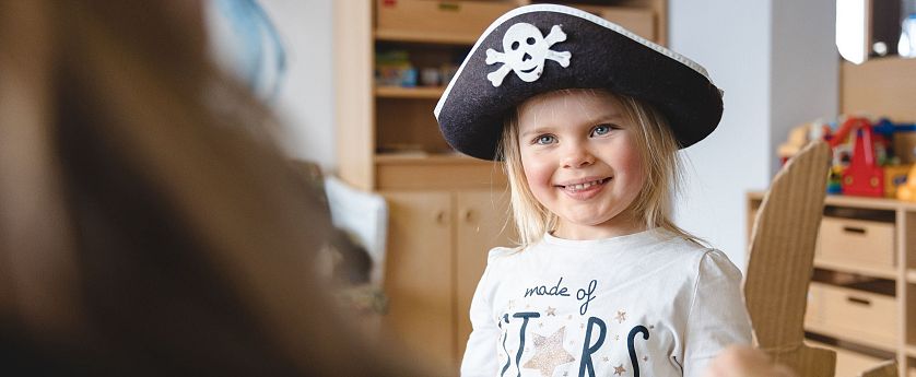 Lächelndes Mädchen mit Piratenhut und Kartonschwert steht in einem Raum mit Holzregalen und Spielzeug.