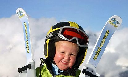 Ein fröhliches Kind steht im Schnee, trägt einen Helm und eine Skibrille. In den Händen hält es kleine Skier mit der Aufschrift "mini-carver". Hintergrund: Berglandschaft.