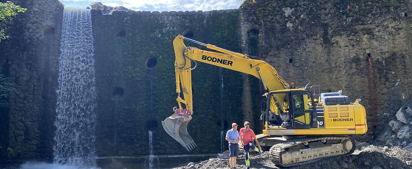 Ein gelber Bagger mit der Aufschrift "BODNER" steht vor einer alten Mauer. Zwei Männer beobachten den Bagger. Im Hintergrund sind große, runde Öffnungen in der Mauer sichtbar.
