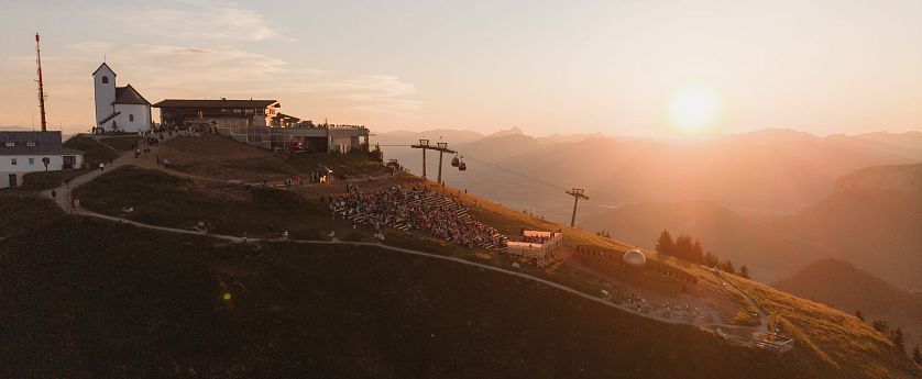 Landschaftsaufnahme der Berge bei Sonnenuntergang mit einer kleinen Berghütte, Lifte und Besucher auf einem Hang.
