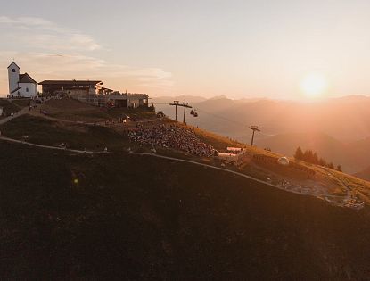 Landschaftsaufnahme der Berge bei Sonnenuntergang mit einer kleinen Berghütte, Lifte und Besucher auf einem Hang.