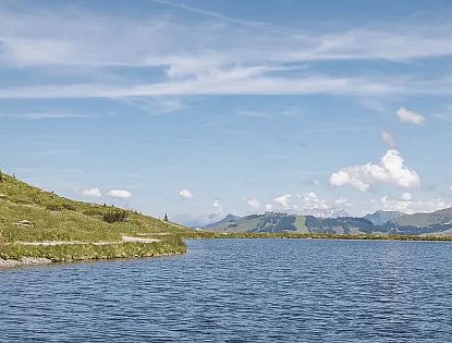 Kristallklarer Bergsee inmitten grüner Almwiesen und Nadelwälder mit Panoramablick auf die Tiroler Alpen an einem sonnigen Sommertag.