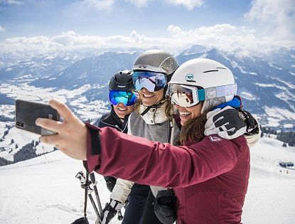 Drei Skifahrerinnen am Ladies Day mit Helmen und Skibrillen machen lachend ein Selfie auf einer verschneiten Piste mit weitem Blick über die winterliche Berglandschaft