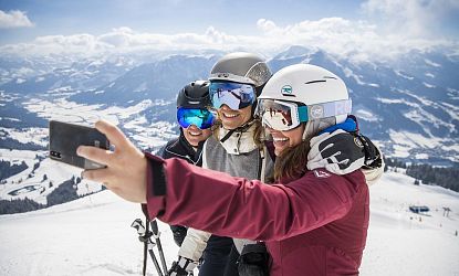 Drei Skifahrerinnen am Ladies Day mit Helmen und Skibrillen machen lachend ein Selfie auf einer verschneiten Piste mit weitem Blick über die winterliche Berglandschaft