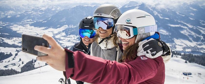 Drei Skifahrerinnen am Ladies Day mit Helmen und Skibrillen machen lachend ein Selfie auf einer verschneiten Piste mit weitem Blick über die winterliche Berglandschaft