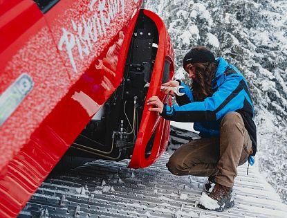 Ein Mann in blauer Jacke prüft das Rad eines roten Fahrzeugs auf schneebedecktem Boden. Winterlandschaft mit Kälte und Schnee im Hintergrund.