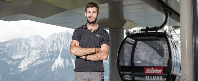 Ein lächelnder Mann in einem schwarzen T-Shirt steht mit verschränkten Armen vor einer Seilbahn. Im Hintergrund sind hohe Berge und ein klarer Himmel zu sehen.